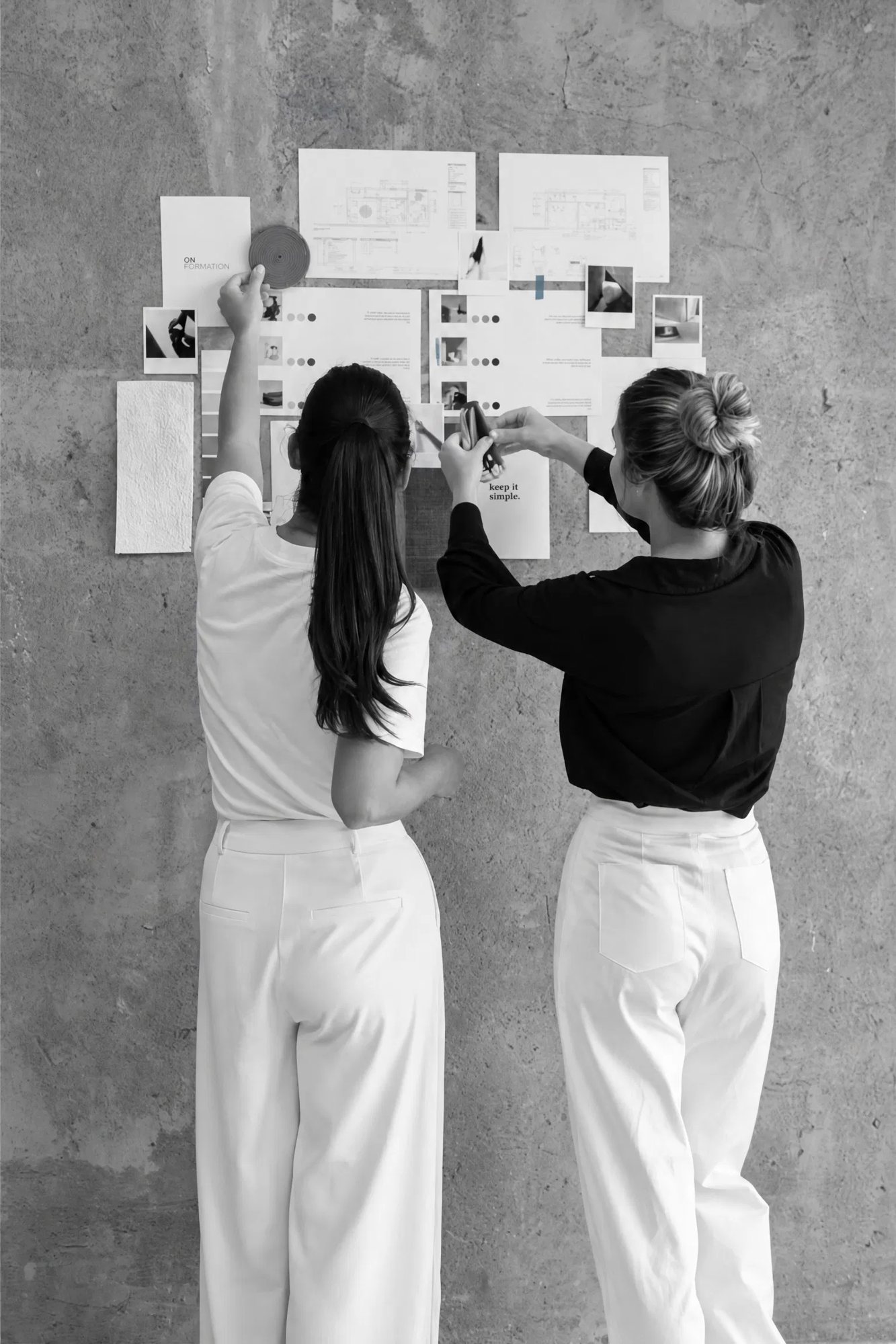 Zwei Frauen ordnen Notizen und Bilder an einer Betonwand, mit KI entworfen